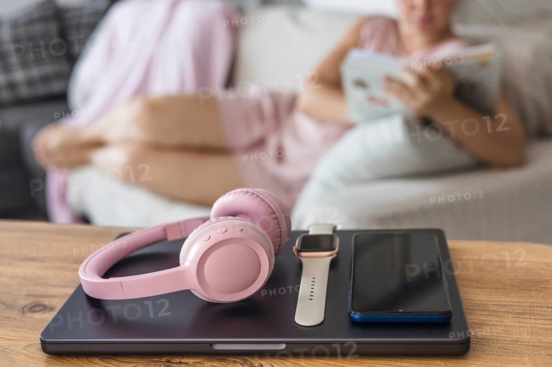 A cozy living room scene with a young woman reading a book. On a wooden table, there are pink headphones, a smartwatch, and a smartphone