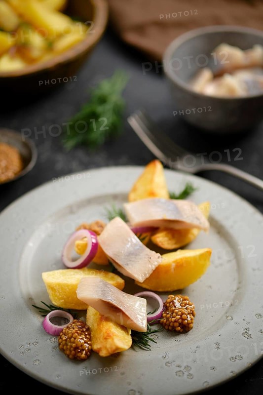 Dish features potato wedges and herring arranged on plate with mustard and sliced onion. This meal appears in a casual indoor dining space with wooden and metallic utensils nearby