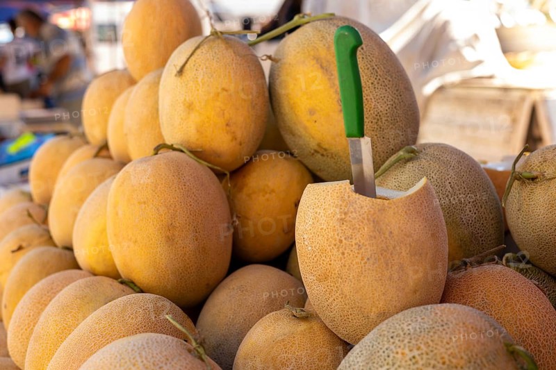Ripe melons stacked at an outdoor market with one melon cut open displaying its flesh