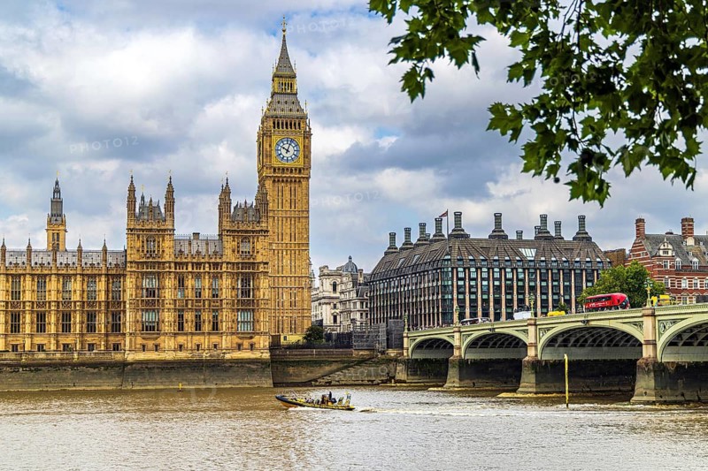 A London bus crosses Westminster Bridge beneath Elizabeth Tower and the Palace of Westminster while a small boat travels the River Thames below. Portcullis House stands beyond the bridge, presenting three eras of Westminster architecture in one riverside view, London, England, United Kingdom
