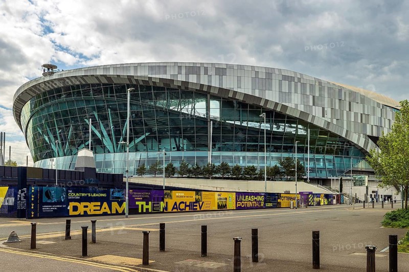 The curved glass and metal facade of Tottenham Hotspur Stadium rises above hoarding on White Hart Lane, London, England, United Kingdom