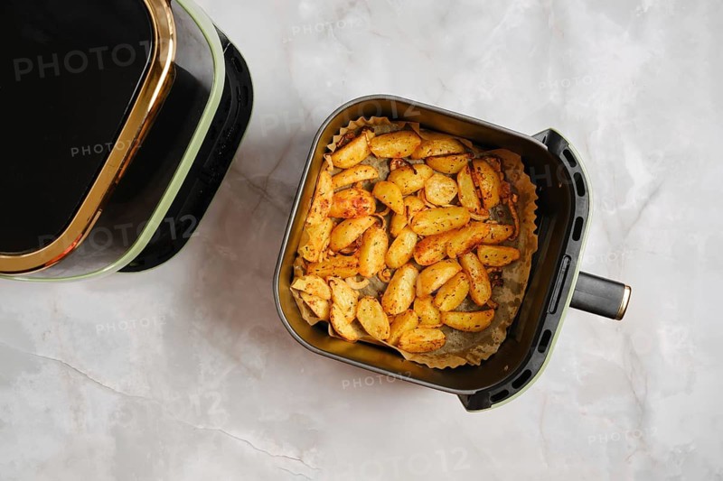 Top view of golden roasted potato wedges cooked in air fryer with spices. Basket sits on marble countertop next to air fryer