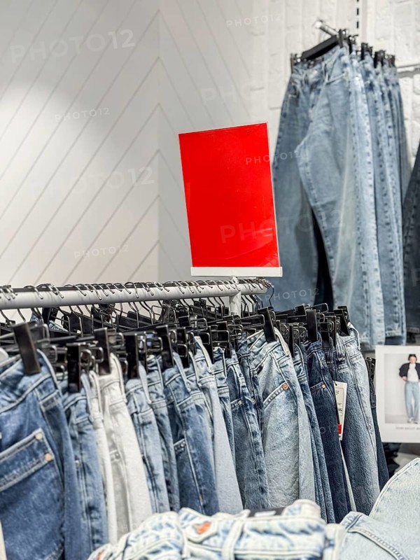 A display of various denim jeans hanging on a rack in a clothing store. A bright red sign is visible in the background, indicating a sale or promotion