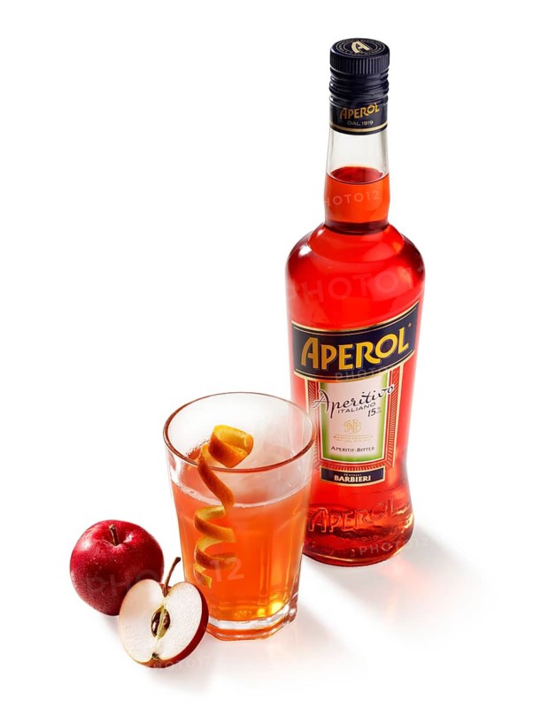 Aperol hot drink, Aperol bottle at the back, apple, half apple and orange zest as garnish, against white background, studio shot