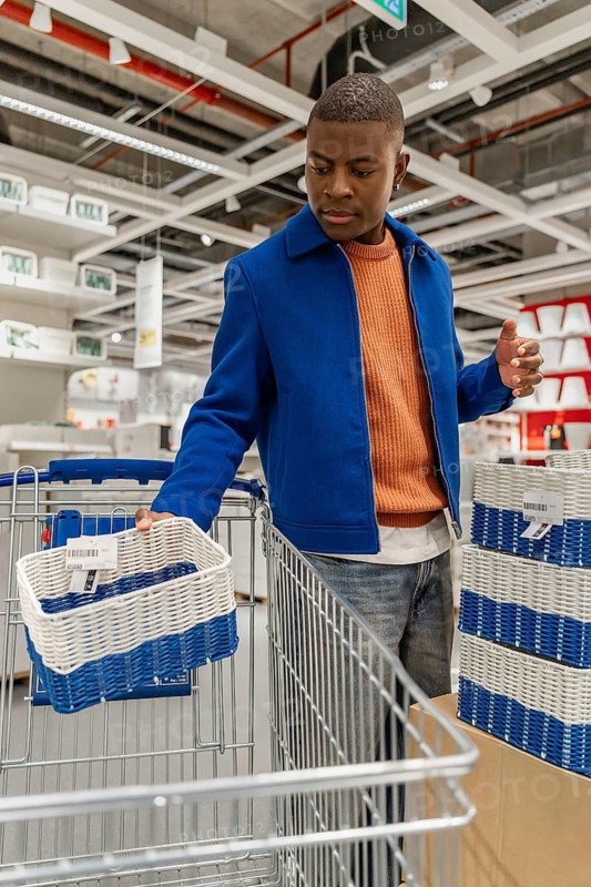 A young Black man with short hair is shopping in a store. He is holding a blue and white shopping basket while looking at items in a cart