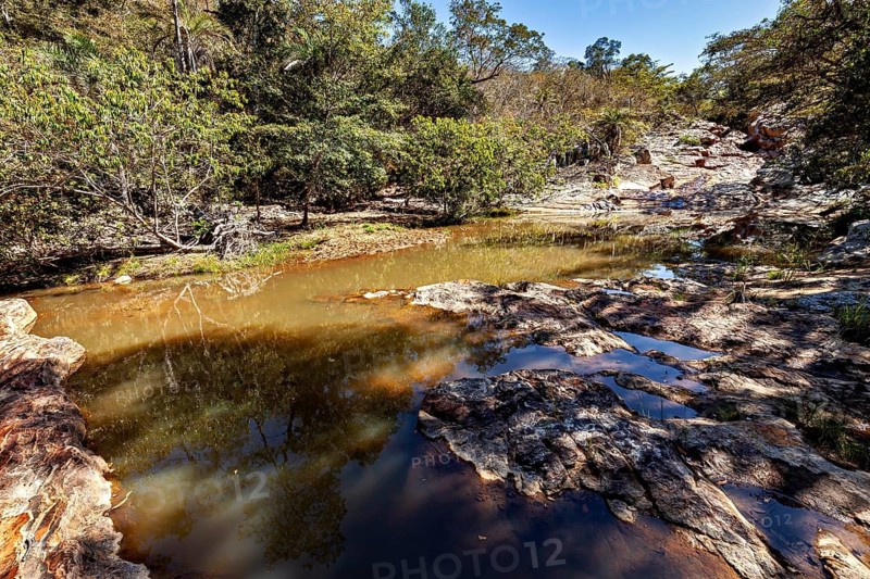 A shallow watercourse snakes through rocks and green vegetation under a blue sky, a small river flows through the thorny savanna of the Caatinga of Bahia in Brazil