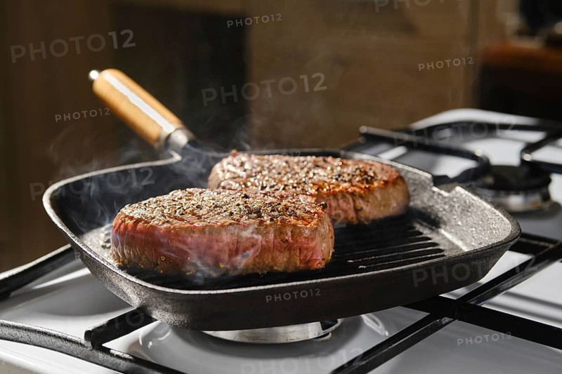 Two beef steaks are cooking in cast iron skillet on stovetop in kitchen. Smoke rises from the sizzling meat. Scene shows preparation of meal