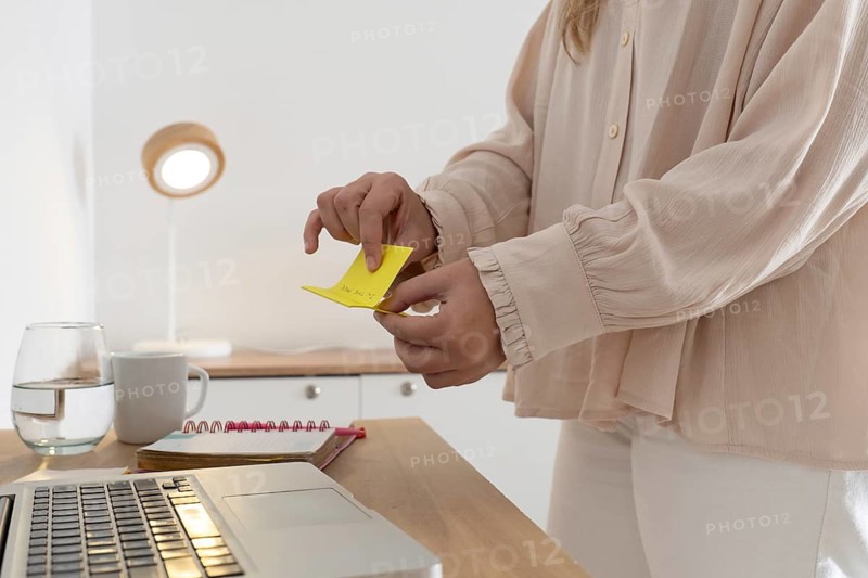 Detailed close up of woman's hands holding and reading a yellow sticky note reminder near laptop, glass of water and mug on wooden desk