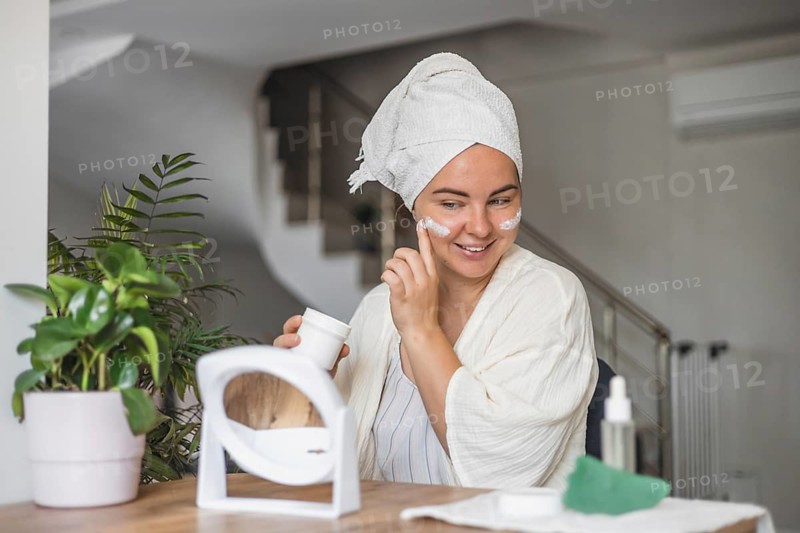 Woman applying skincare while enjoying a beverage in a cozy indoor setting in the morning