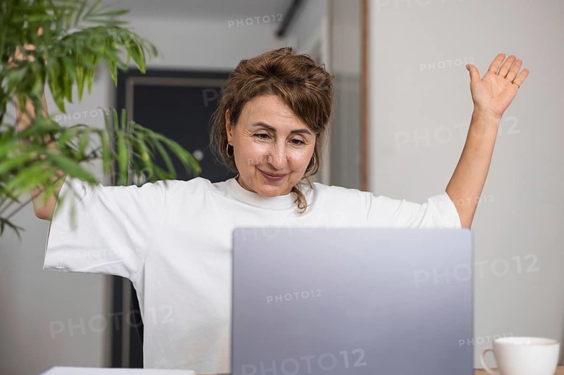 Portrait of happy smiling successful mature investor woman in white clothes, celebrating profit, retirement savings, making winner hand, sitting at laptop at home, green flower in pot on the table