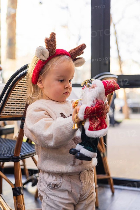 A young Caucasian girl with blonde hair wears reindeer antlers. She holds a Santa Claus figurine, surrounded by a festive atmosphere in a cozy cafe