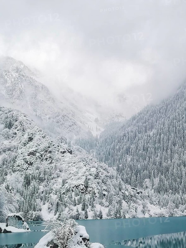 Ice-covered turquoise water of a mountain lake against the backdrop of mountains in fog and clouds. Winter landscape, Snow-covered mountains and pine trees surrounding a calm blue lake under a misty sky, Lake Issyk, Kazakhstan