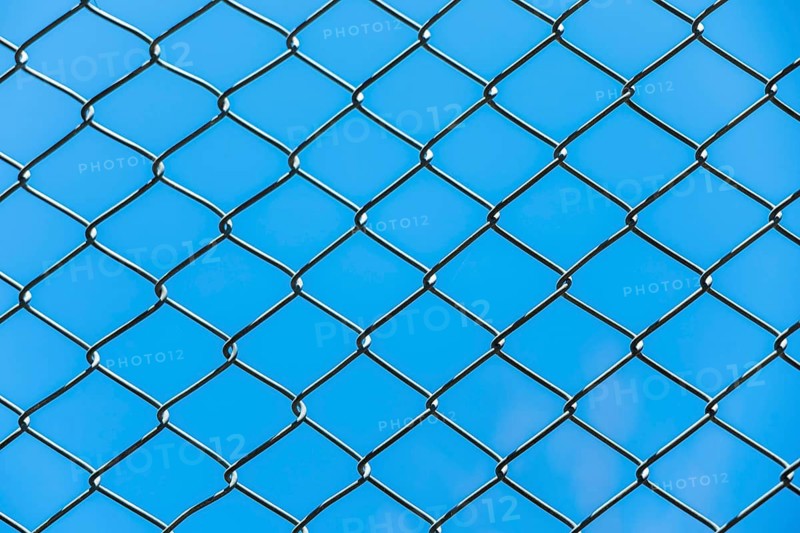 A close-up view of a chain-link fence against a clear blue sky. The fence features a diamond pattern with metallic wires creating a textured background