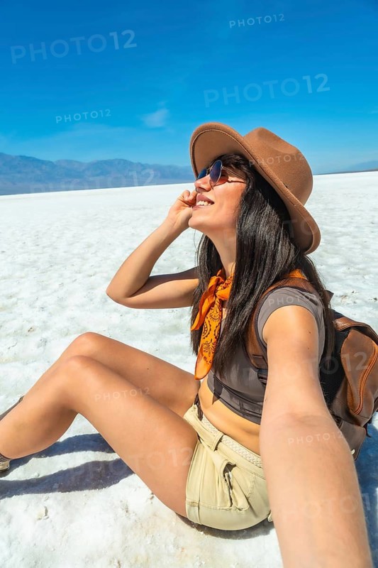 Young woman smiling in sunglasses and a cowboy hat, taking a selfie while relaxing on the vast white salt flats of badwater basin, death valley under clear blue sky