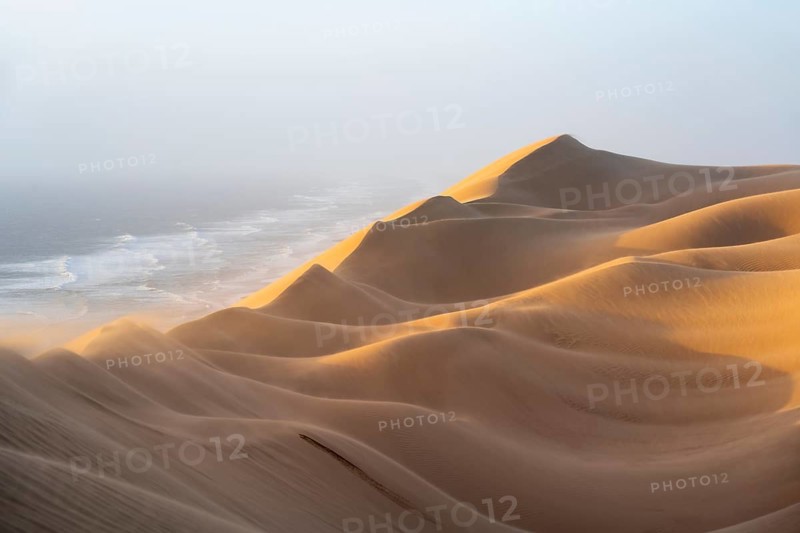 Sand dunes and sea in evening light, wind blowing sand across the dunes, view of the Atlantic coast from high sand dunes, Sandwich Harbour, Namib Naukluft Park, Namibia