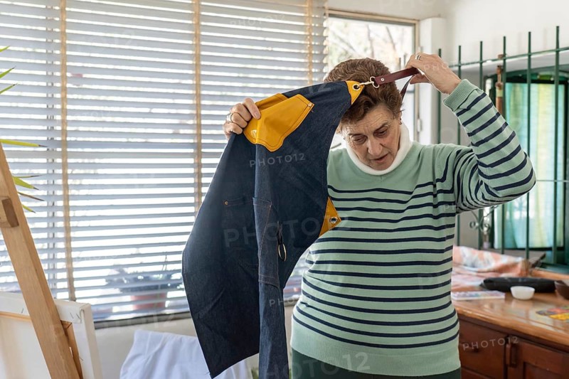 Older woman lifts a blue denim apron with yellow detail over her head to remove it, standing near a wooden easel after finishing her painting session