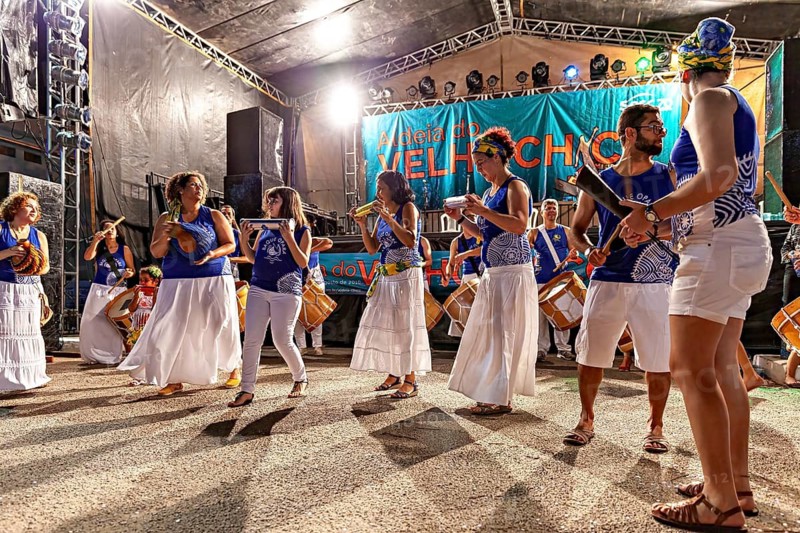 Women in white skirts and blue tops dance to drum music at a nighttime show, a band plays and dances at a festival in Petrolina in Brazil