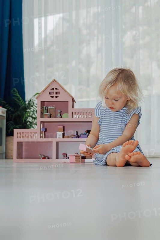 Child toddler blonde girl in blue dress playing with a wooden pink toy house at home