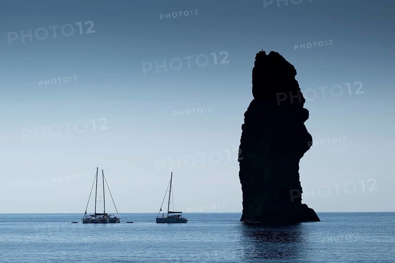 Silhouette of a towering monumental rock pin, sailboats in the calm sea, near Filicudi, Aeolian or Lipari Islands, Southern Italy, Italy