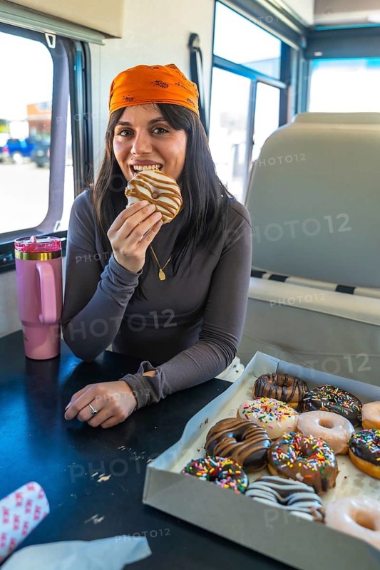 Young woman wearing a bandana and long sleeve shirt biting into a glazed donut from a large box of assorted pastries while traveling in a recreational vehicle