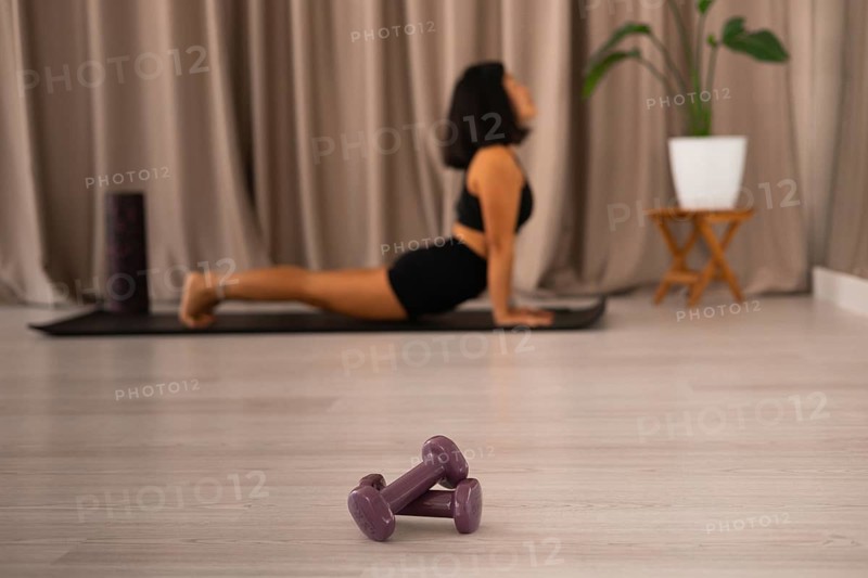 Woman performing yoga pose on mat, dumbbells positioned in front, surrounded by calm setting