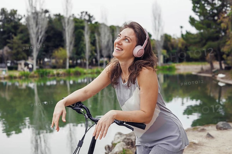 Young Caucasian woman with brown hair and pink headphones smiles while leaning on an electric scooter by a serene lake in a park