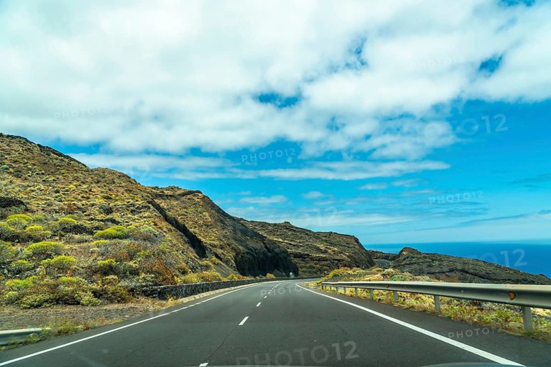 Road tripping along a coastal highway in el hierro, canary islands, showing a winding asphalt road with volcanic hillsides and the atlantic ocean under a blue sky with white clouds
