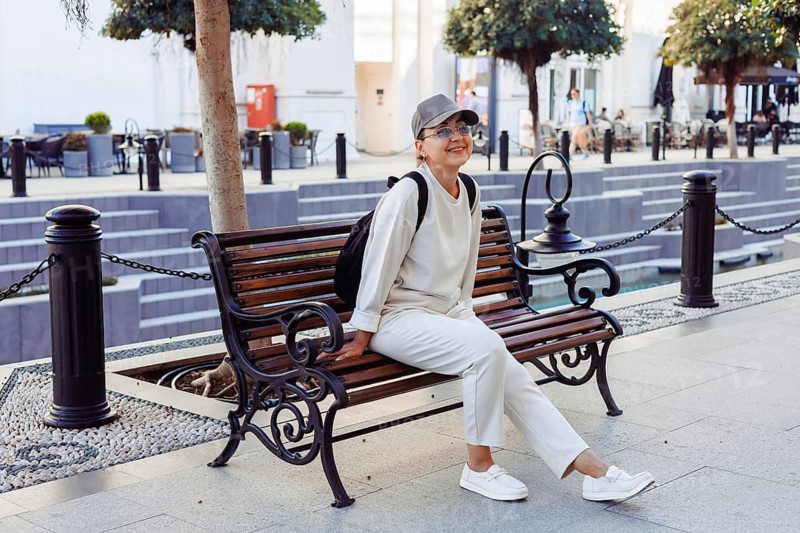 A cheerful middle-afed caucasian woman sits on a wooden bench, enjoying a sunny day in an outdoor area filled with trees and people. Shopping open air center, mall