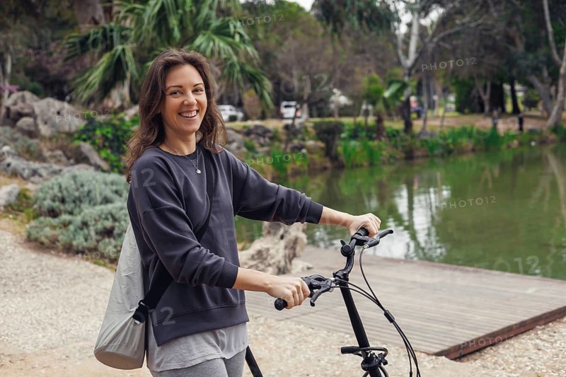 Smiling young Caucasian woman with brown hair stands beside a bicycle near a serene lake in a park. Green trees and rocks are visible in the background
