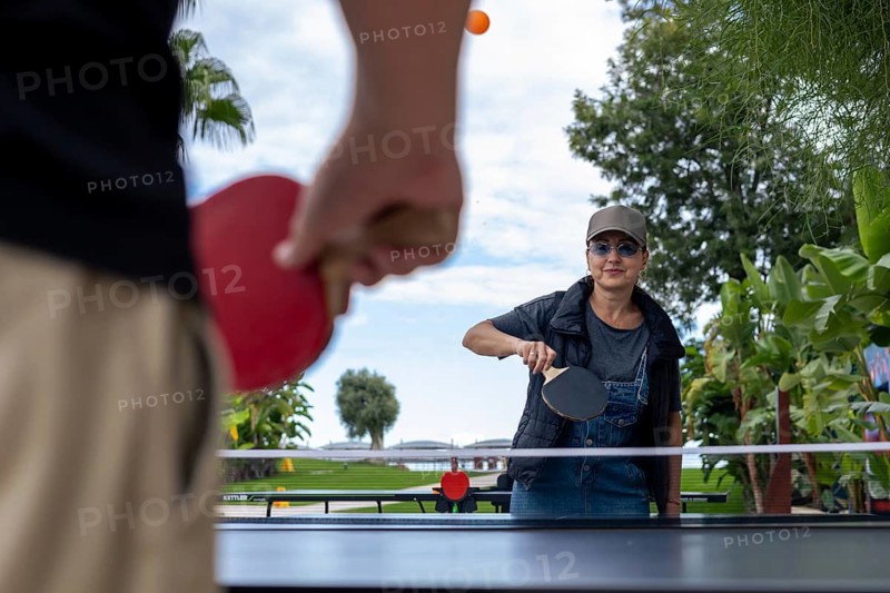 Guests engage in a friendly game of table tennis. The vibrant atmosphere showcases palm trees and clear sky, creating a perfect day for activities. Ping pong game outdoor, hobby and leisure activity