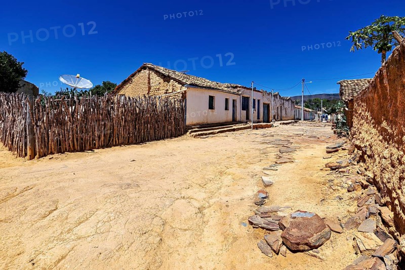 A sandy path leads past blue and white houses and a rustic wooden fence, settlement and villages in the Caatinga of Pernambuco and Bahia in Brazil