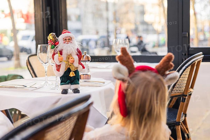 A young girl with blonde hair wearing reindeer antlers sits at a table. Christmas decorations include Santa figurines and a festive atmosphere