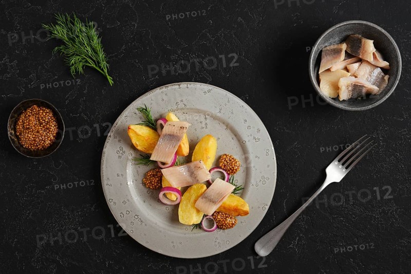 Plate with potato wedges arranged beside pieces of herring and decorated with mustard and onion. There is small bowl with more herring nearby. Green herbs add color to the setting