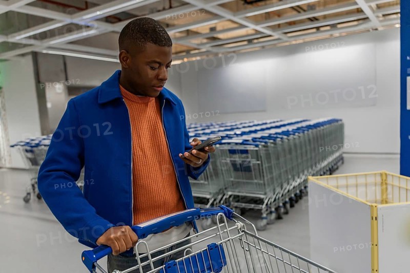 A young Black man in a blue jacket and orange shirt checks his phone while pushing a shopping cart in a spacious store aisle filled with carts