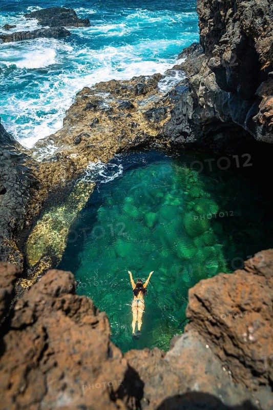 Woman swimming on her back in the clear turquoise waters of a natural volcanic pool, surrounded by rugged coastal rocks with the ocean waves breaking nearby during a sunny day