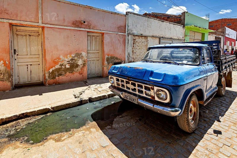 A blue pickup truck stands in front of old, dilapidated buildings on a sunny paved street, street scene in Campo Formoso in Caatinga in Bahia in Brazil