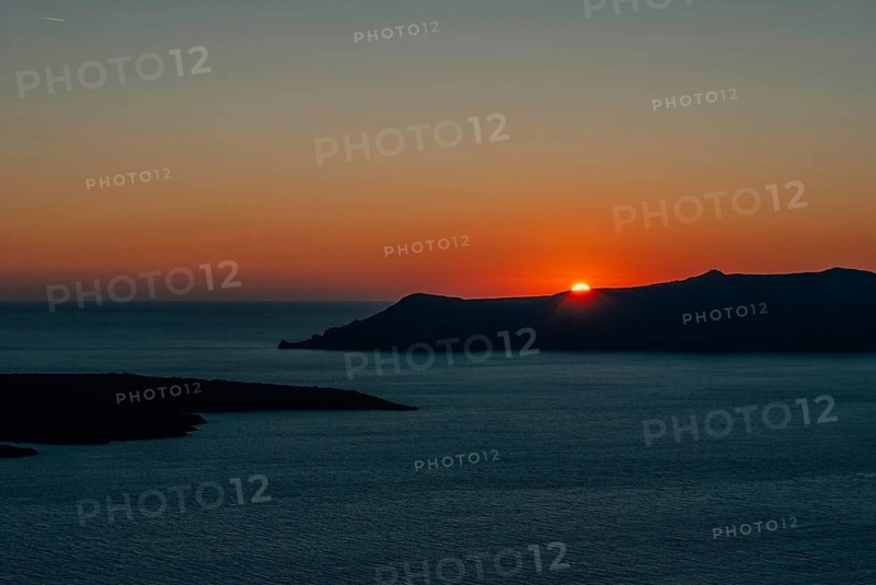 The sun at sunset almost disappeared behind the island, Santorini, Greece, A tranquil sunset over the ocean with a silhouette of a distant land against an orange and blue sky, Santorini, Greece