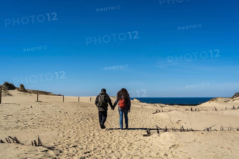 A couple of travelers with a backpack, hand in hand, walks along the sandy Dead Dunes, on the Curonian Spit, on the shore of the Baltic Sea, A couple walks hand in hand along a sandy path with dunes and the sea in the background under a clear blue sky, Curonian Spit, Neringa, Lithuania