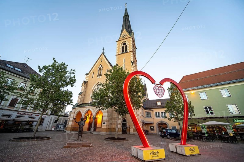 Illuminated St. Nicholas Church and bronze figure of Francis of Assisi, blue hour, heart at Paul Watzlawick Square, Villach, Carinthia, Austria
