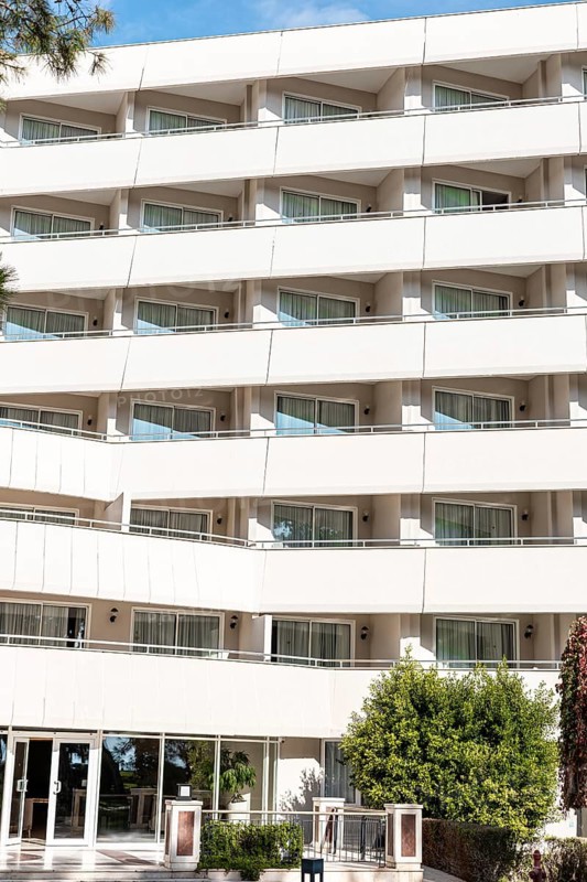 A modern hotel building with multiple balconies and large windows. The structure features a clean, white facade and is surrounded by greenery