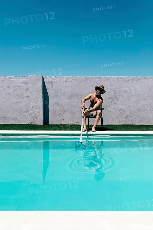 A shirtless man in shorts and a hat cleans a swimming pool with a long net skimmer under a bright blue sky. He sits on a chair near a concrete wall, embodying a summer vibe