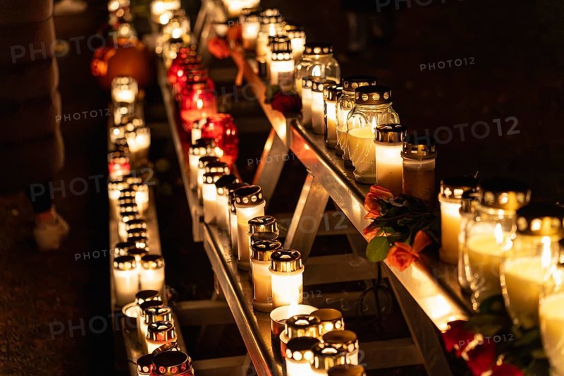 Lit candles in glass holders glow on a multi tier metal structure at night, with red and orange roses placed among them. The scene was photographed at Skogskyrkogarden cemetery in Stockholm during All Saints Day and captures a quiet moment of remembrance and reflection