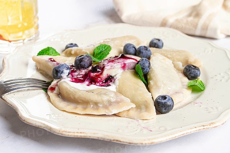 Handmade dumplings filled with sweet ingredients, arranged beautifully on a white plate, topped with berry sauce, fresh blueberries, and mint leaves, ready to enjoy