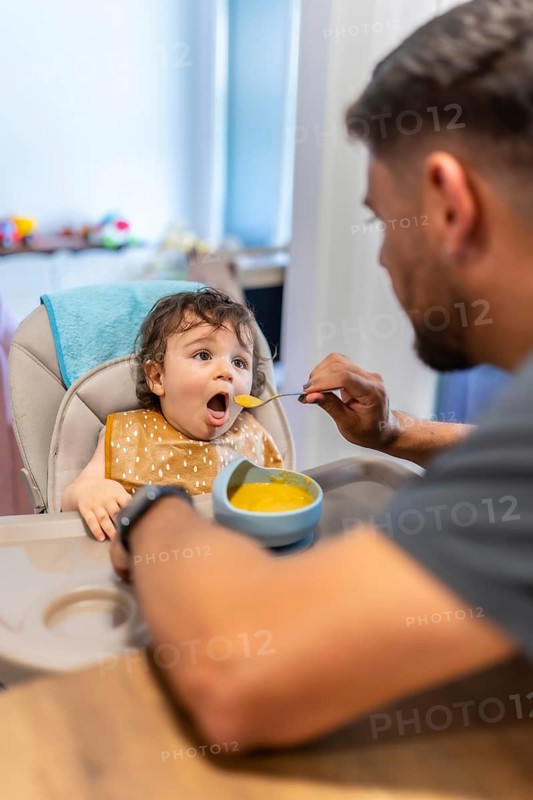 Young father lovingly feeding his adorable baby son nutritious vegetable puree while sitting in a modern high chair, celebrating healthy eating habits and family bonding during mealtime at home