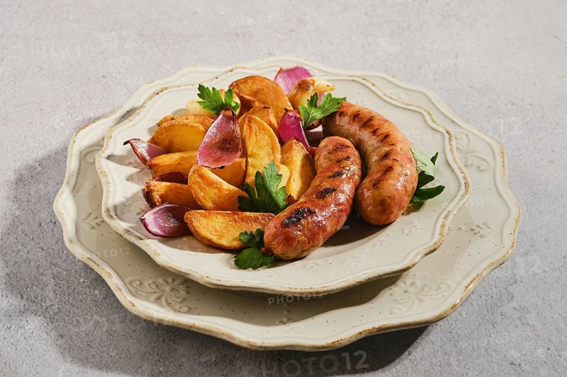 Sausages cooked on grill served with golden roasted potato wedges and purple onions on cream two-tier plate. Green herbs add color and flavor. Enjoyed during lunch or dinner at home