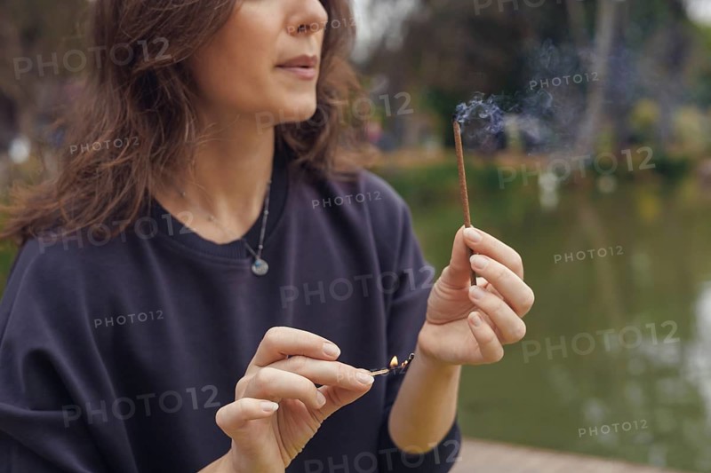A young Caucasian woman with long brown hair sits by a pond, holding an incense stick. Smoke rises gently from the stick, creating a serene atmosphere