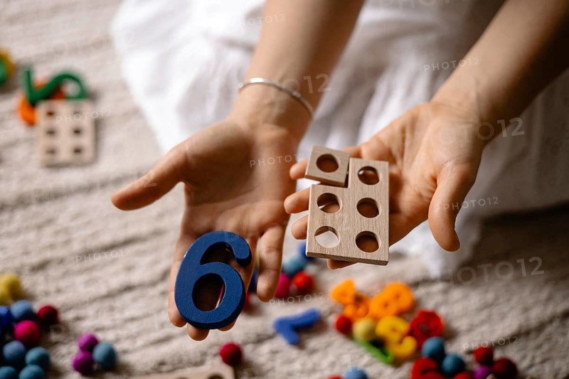 Hands holding a blue number six and wooden toy with colorful scattered toys