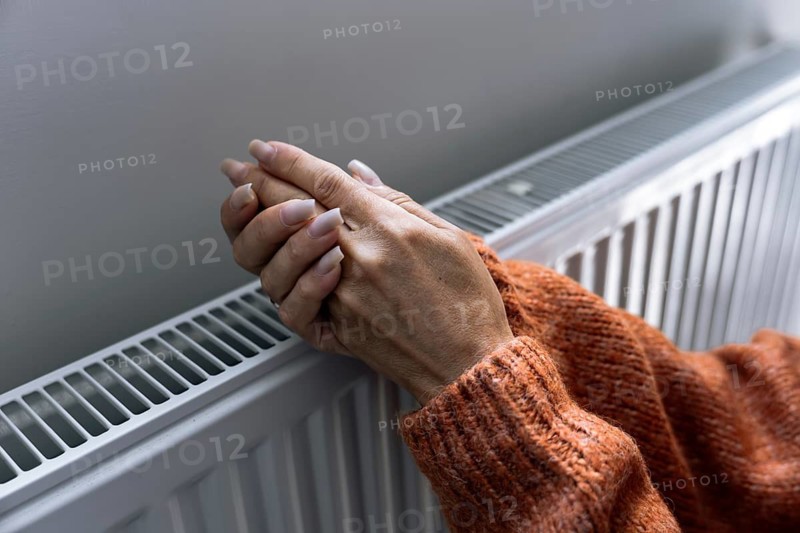 Woman warming hands near home radiator during heating season in Europe. Concept of rising utility bills, expensive household heating, energy crisis, winter budgeting and cost of living.