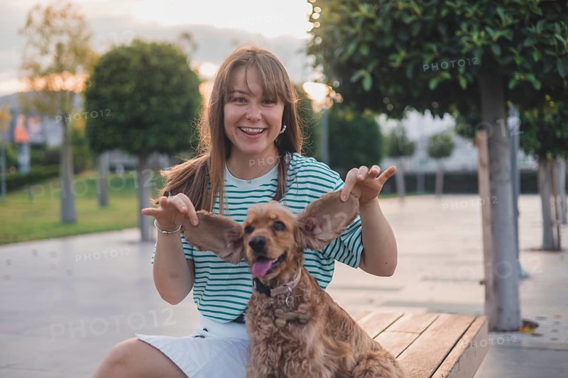 A woman playfully holds a dog's ears while sitting on a bench in a sunlit park