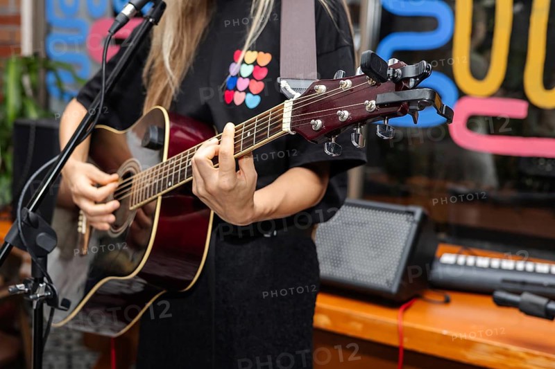 A young woman with long blonde hair plays an acoustic guitar on stage. Colorful decorations are visible in the background, creating a lively atmosphere